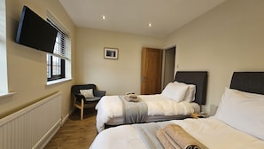 A hotel room with two beds, a chair, a TV mounted on the wall, and a window with blinds. - Seashells Cottage (Lytham St. Anne's)