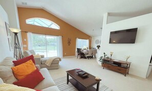 A spacious living room with a high ceiling, a flat-screen TV mounted on the wall, a dining area with a table and chairs, and a large window providing natural light. - 480-Beautiful 3-Bed Villa w/ SE-Facing Pool & Conservation View (Davenport)