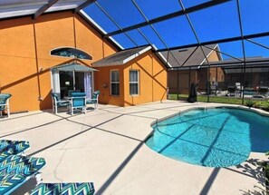 A swimming pool with a covered patio area, a small seating area with chairs, and a clear blue sky.