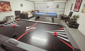 A pool table with a red and white design, a blue pool table, and a sofa in a room with white walls and posters.