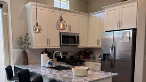 A modern kitchen with white cabinets, a stainless steel refrigerator, a built-in microwave, and a granite island with bar stools. - Desert Olive Retreat - Sleeps 4- 5 min fr Verrado Marketplace 30 min fr airport (Buckeye)