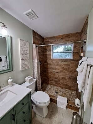 A bathroom with a shower, a toilet, a sink, and a mirror. There is a window above the shower and a sign with house rules on the wall.