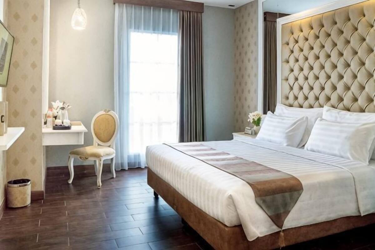 Habitación de hotel con una cama grande, un escritorio con silla, una ventana con cortinas y una lámpara fijada a la pared.