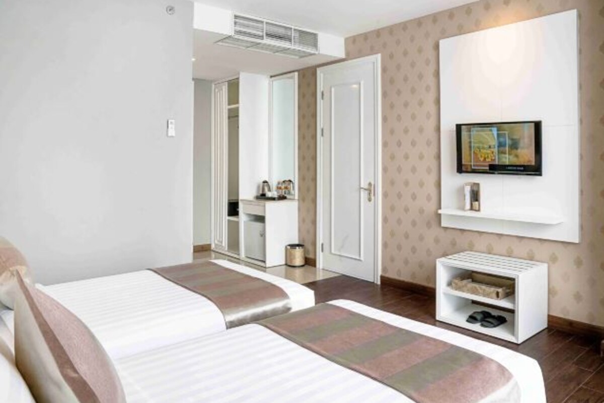 A hotel room with two beds, a TV mounted on the wall, a wardrobe, and a small shelf with items.