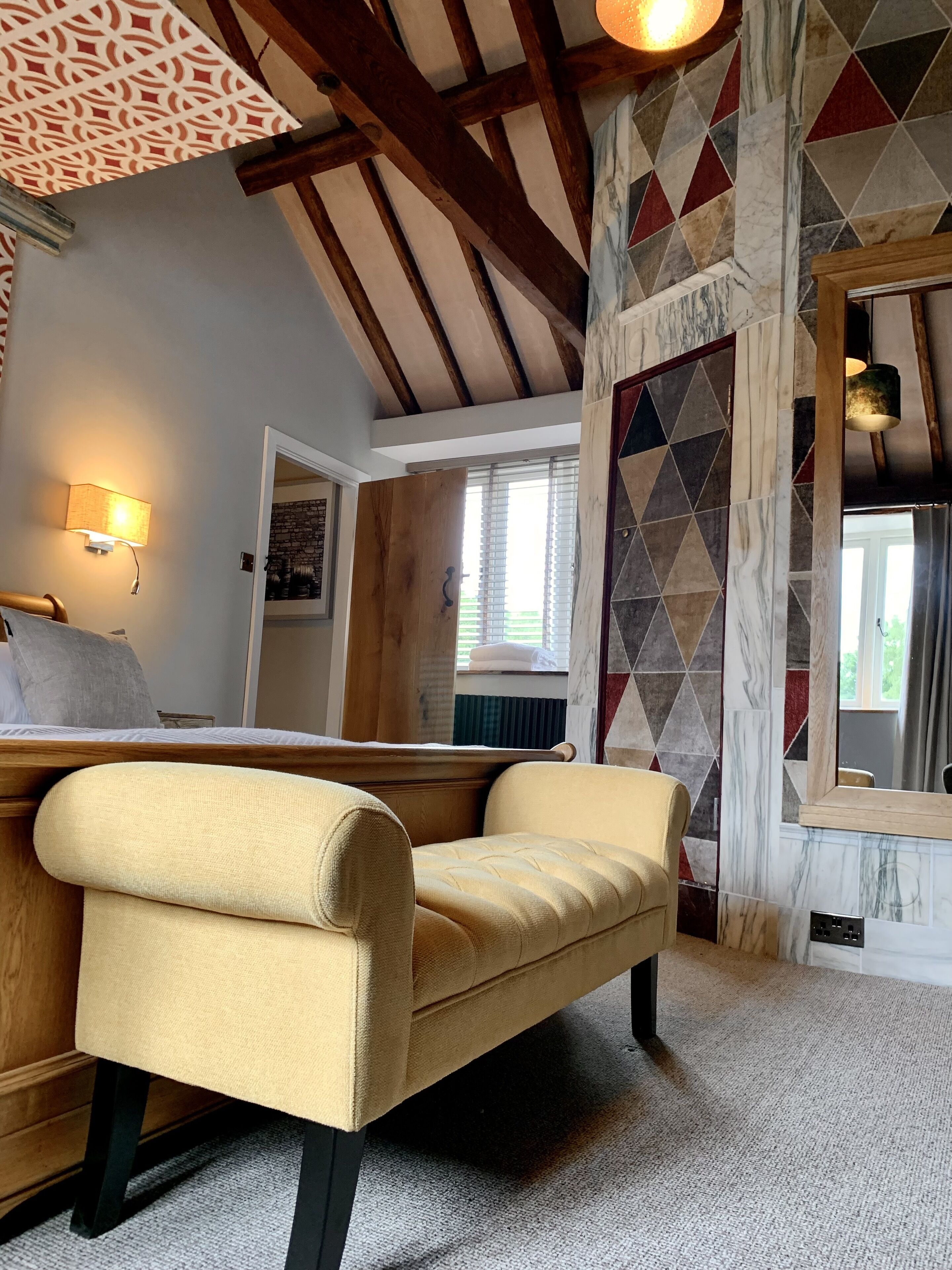 A cozy room with a wooden ceiling, a bed with a headboard, a beige armchair, and a patterned wall.