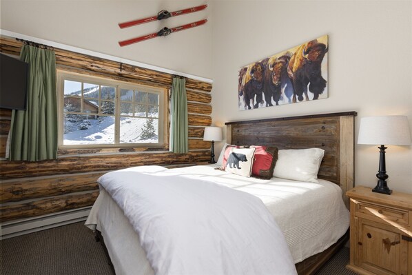 A cozy room with a large bed, a wooden headboard, a window with a view of a snowy landscape, and a painting of bison on the wall. - Moonlight Basin True Ski-in/ski-out Cowboy Heaven Cabin, Hot Tub, Views (Big Sky)