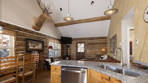 A rustic cabin interior with a kitchen featuring granite countertops, wooden cabinetry, and a stainless steel dishwasher. A mounted elk head decorates the wall above the kitchen counter.