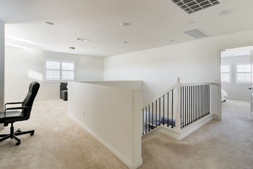 Ein geräumiges, helles Zimmer mit Teppichboden, einem schwarzen Bürostuhl, einer weißen Treppe mit schwarzer Geländer und einem Fenster mit Jalousien.