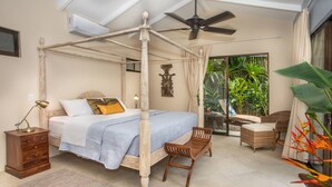 Una cama con dosel, una mesita de noche con lámpara, una silla y vistas a un jardín tropical a través de un ventanal.