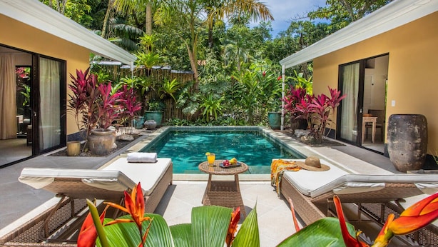 Un área de piscina privada con sillones de descanso y una mesa, rodeada de exuberante vegetación tropical y una piscina cristalina.