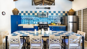 Una cocina con armarios azules, un microondas, una estufa y un refrigerador. Hay una mesa a cuadros con sillas y un reloj en la pared.