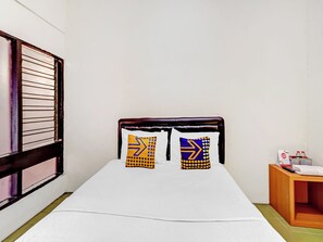 A single bed with a white mattress and two pillows with geometric patterns, a wooden headboard, a wooden nightstand with a remote control, and a window with blinds.