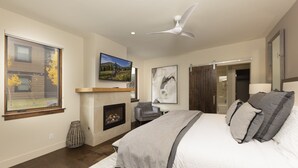 Une chambre avec une cheminée, un ventilateur de plafond, un lit avec des oreillers, un téléviseur fixé au mur et une fenêtre donnant sur une maison et des arbres.