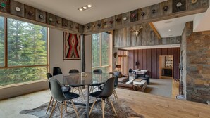 A spacious room with a large glass table surrounded by chairs, a stone wall, and a view of a forest through large windows. - Mod Haus by Tahoe Getaways - West Shore w/ Private Beach Access & Pier + Close to Lake! (Homewood)