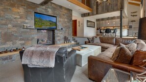 A modern living room with a stone wall, a fireplace, a large sofa, and a television mounted on the wall. - Mod Haus by Tahoe Getaways - West Shore w/ Private Beach Access & Pier + Close to Lake! (Homewood)