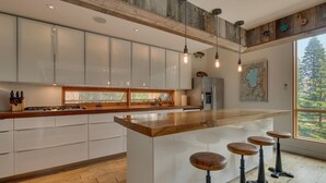 A modern kitchen with a large island, wooden stools, and a view of the forest through the window. - Mod Haus by Tahoe Getaways - West Shore w/ Private Beach Access & Pier + Close to Lake! (Homewood)