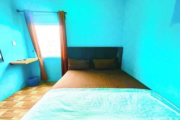 A small, narrow room with a bed, a window with curtains, a small shelf, and a blue bucket on the floor.