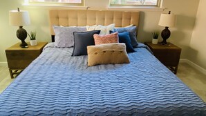 Une chambre à coucher avec une tête de lit capitonnée, deux tables de chevet avec des lampes, un lit recouvert d’une couvre-lit à motifs bleus, et deux fenêtres donnant sur la forêt.