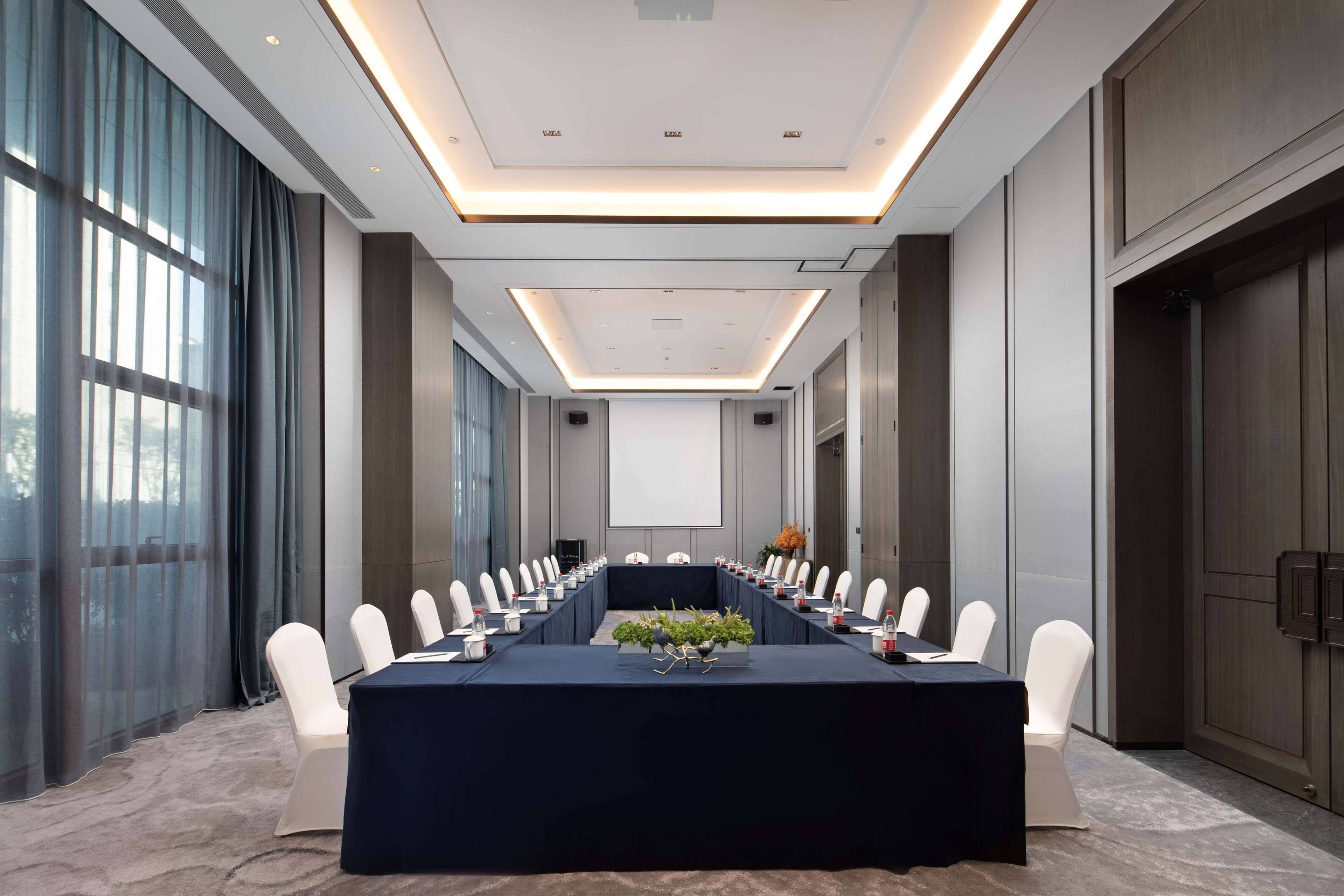 Un salón de conferencias con una larga mesa, sillas blancas y una pantalla de proyector.