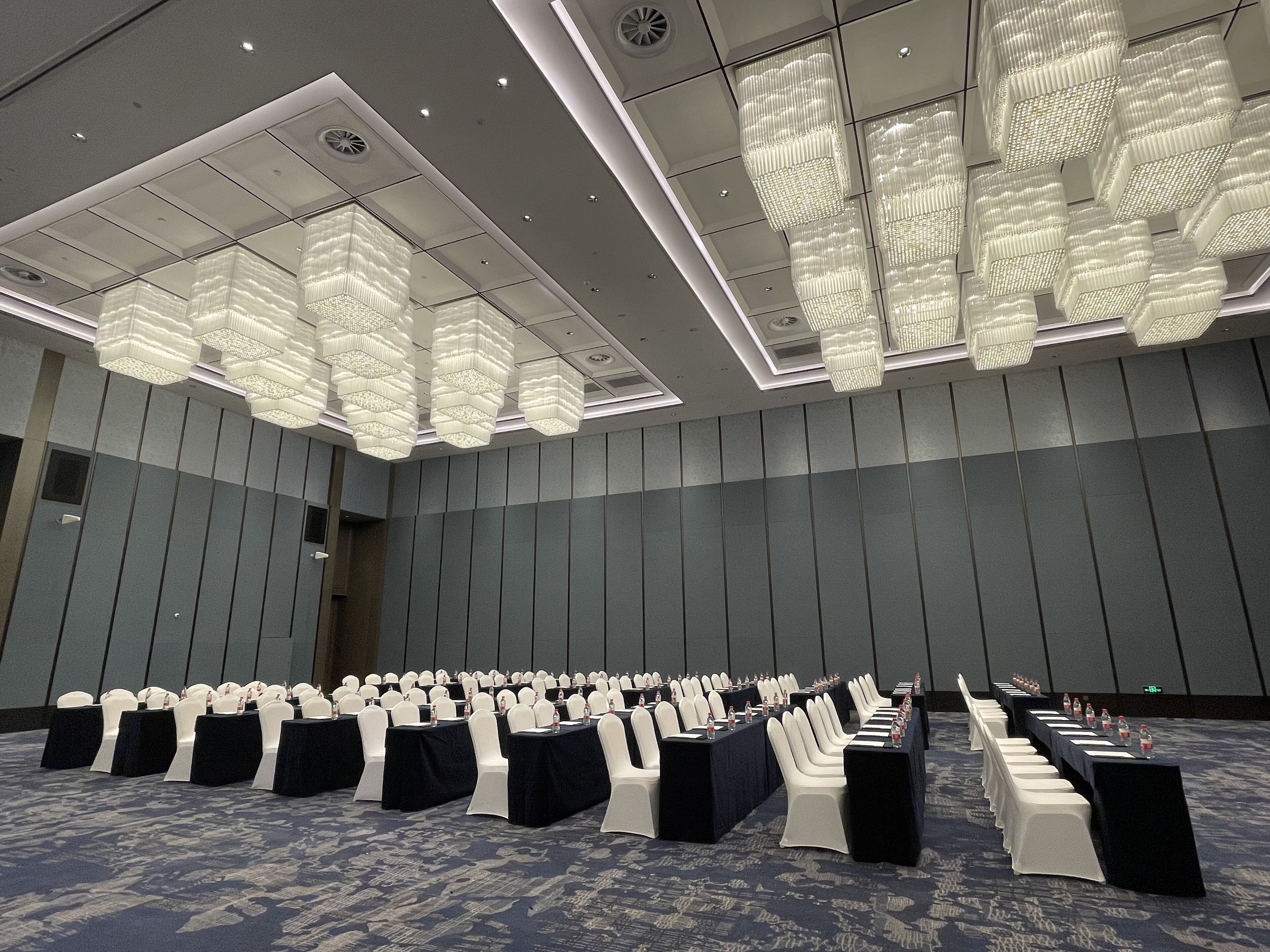 Un salón de conferencias con filas de mesas y sillas, grandes candelabros colgantes y una alfombra estampada.