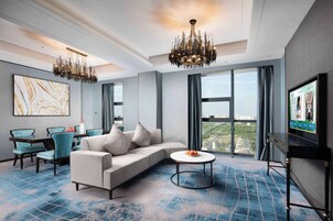 A modern hotel room with a large sofa, a round coffee table, and a television. There is a large window offering a city view.
