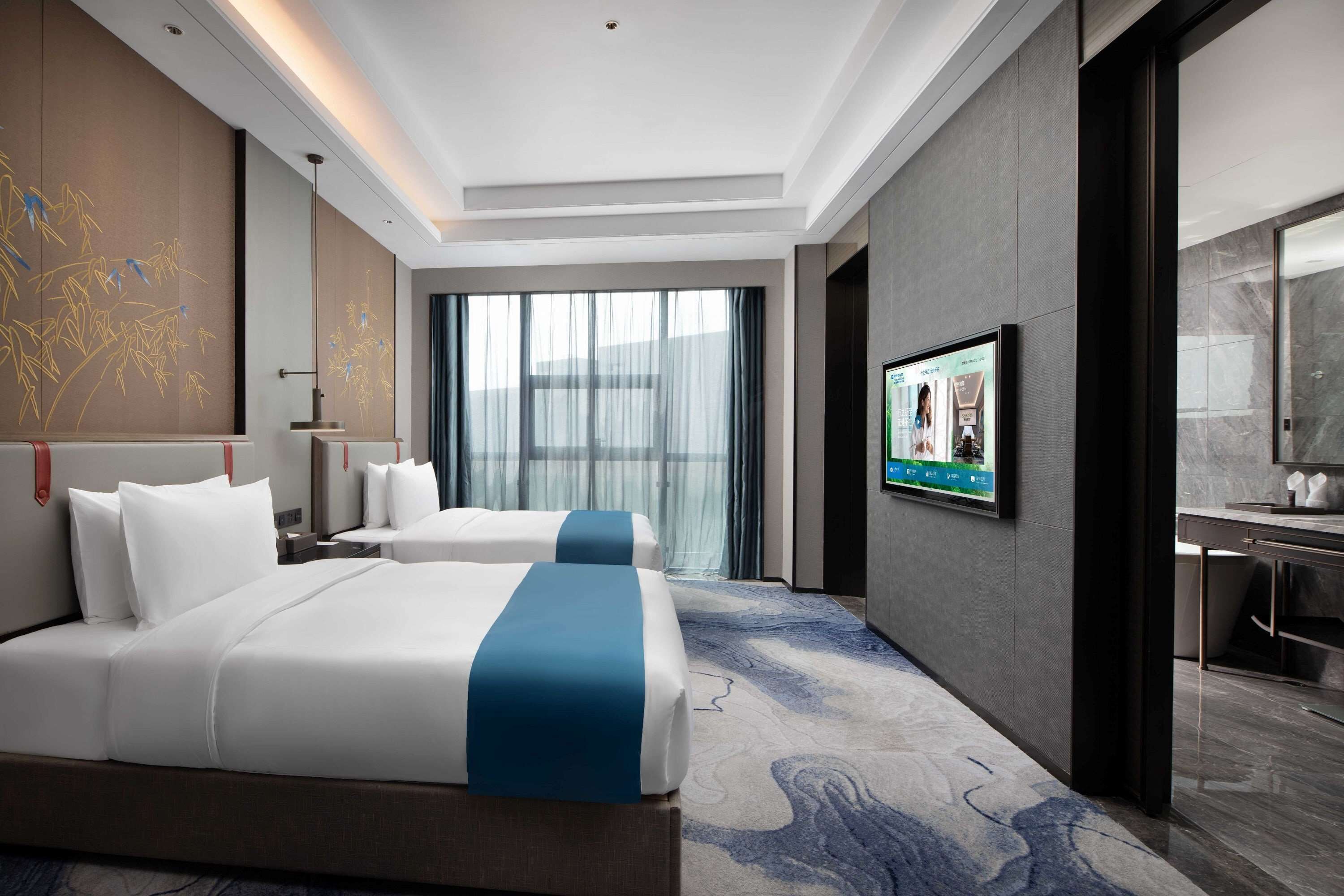 A hotel room with two beds, a large window with curtains, a flat-screen TV, and a modern bathroom.