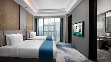 A hotel room with two beds, a large window with curtains, a flat-screen TV, and a modern bathroom.