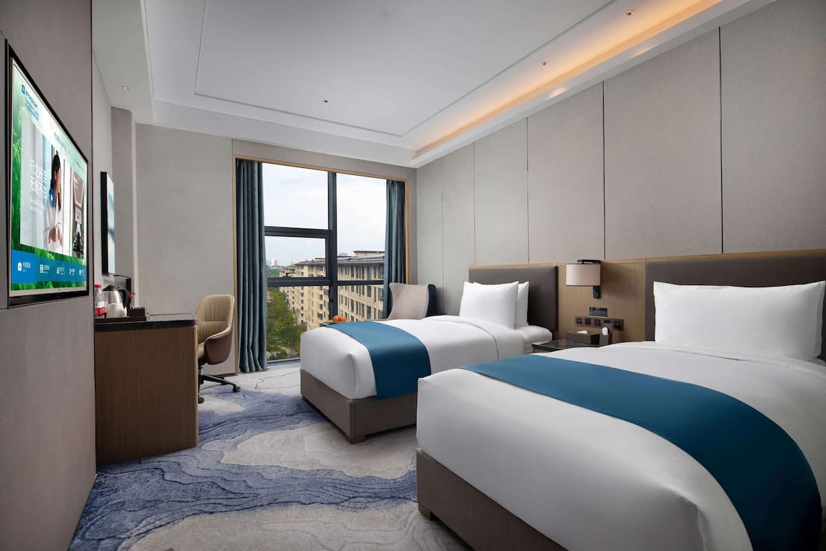 Habitación de hotel con dos camas, un escritorio, una silla, un televisor y un ventanal grande con vistas a la ciudad.