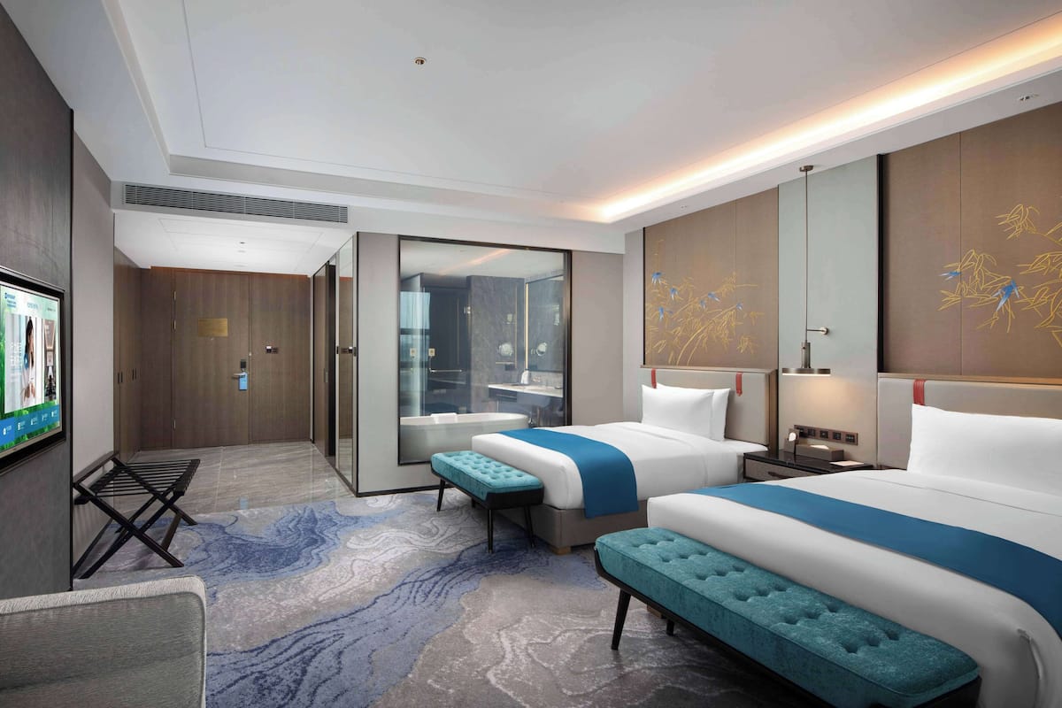 Habitación de hotel moderna con dos camas, una zona de estar, televisor y baño visible a través de una puerta de cristal.
