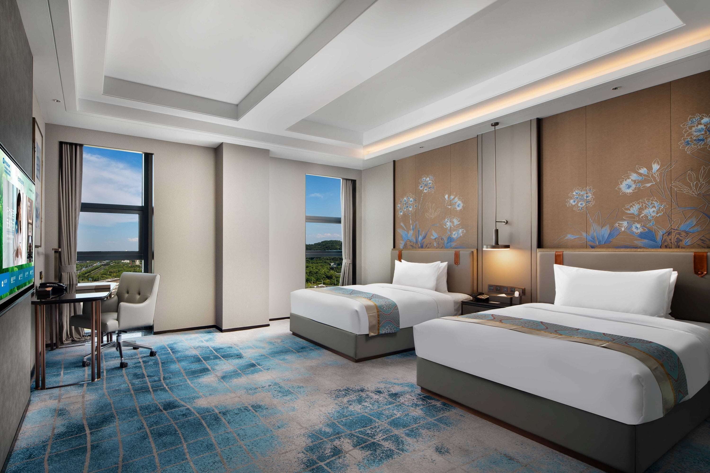 Une chambre d’hôtel avec deux lits, un bureau et une chaise. De grandes fenêtres offrent une vue sur l’extérieur.