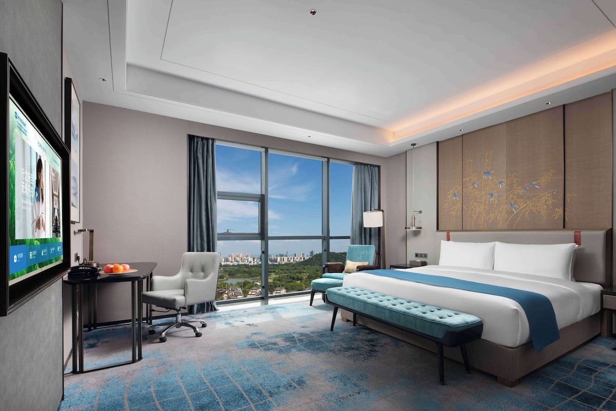 Habitación de hotel con una cama grande, un escritorio con una silla, un televisor y vistas a un paisaje urbano a través de amplios ventanales.