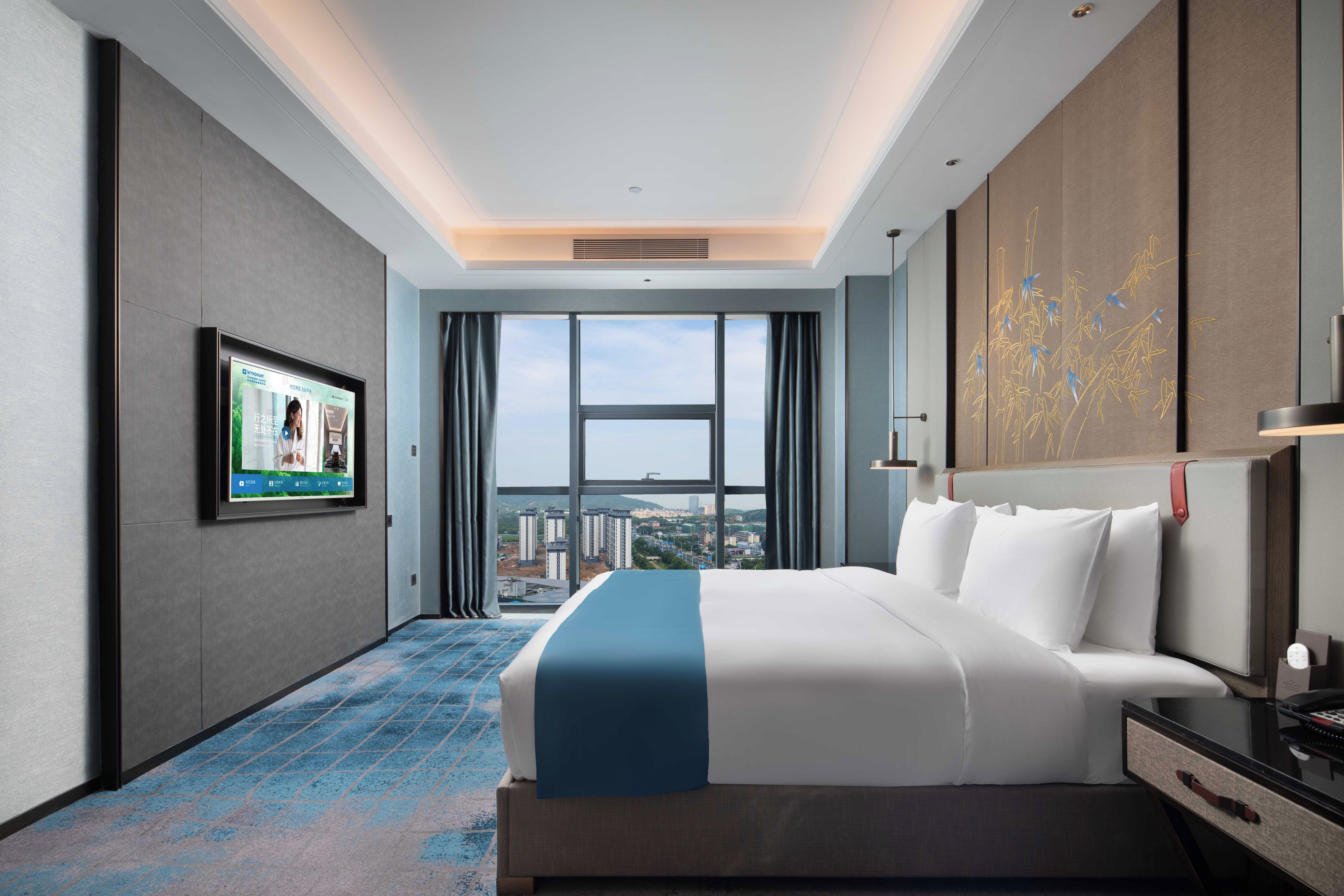 A hotel room with a large bed, a television on the wall, and a view of buildings through the window.