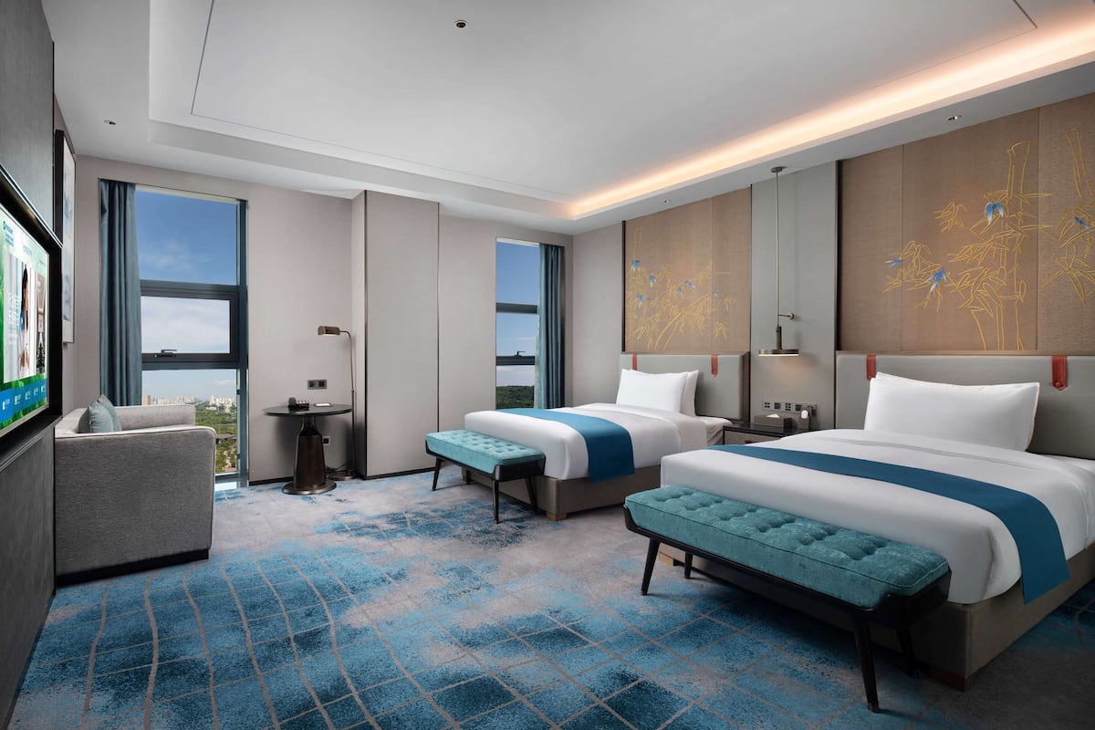 Habitación de hotel con dos camas, un sofá, una mesita y un ventanal amplio con vistas a la ciudad.