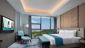 Habitación de hotel con una cama grande, un televisor en la pared, una silla, un banco y vistas de la ciudad a través de amplias ventanas.