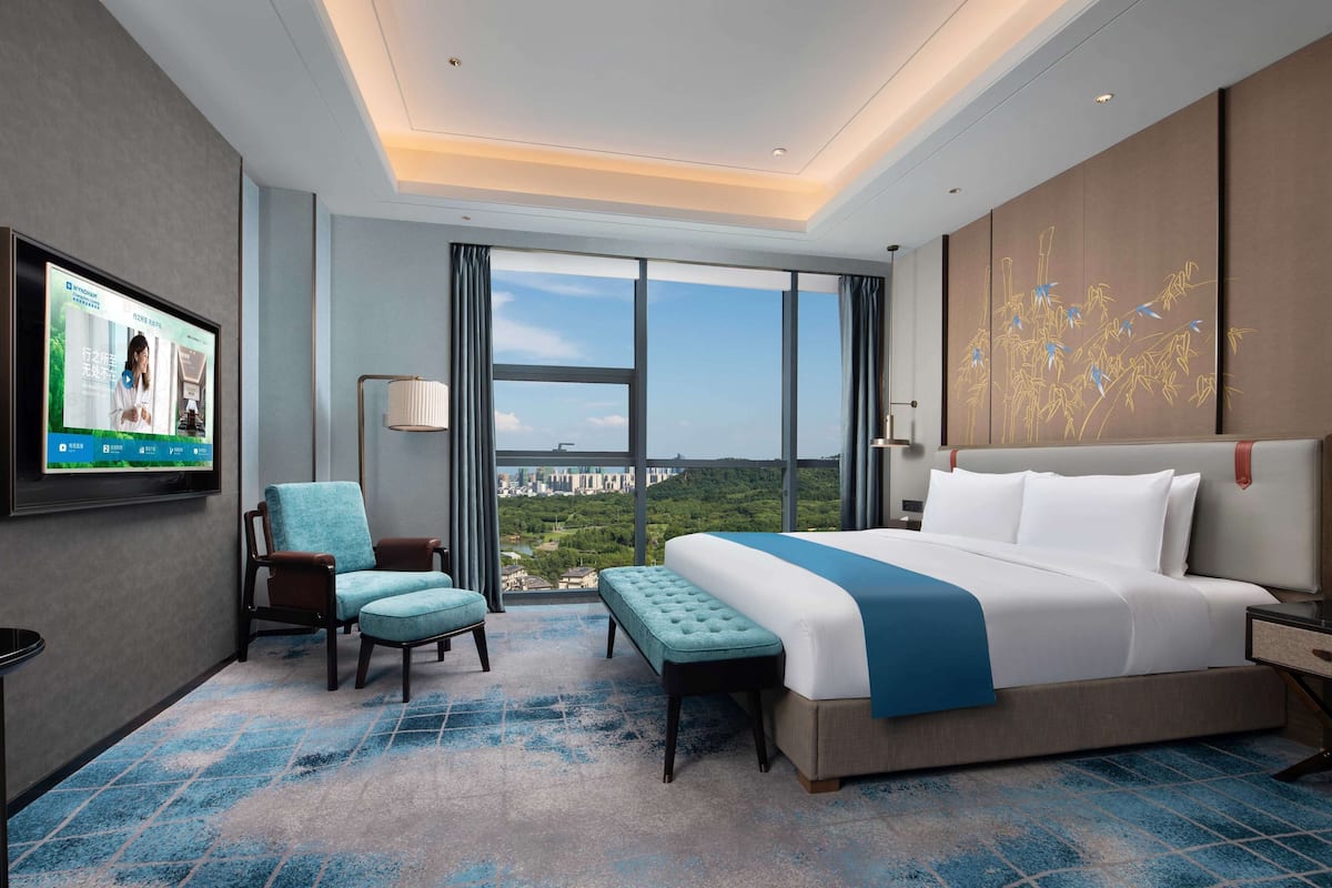 Habitación de hotel con una cama grande, un televisor en la pared, una silla, un banco y vistas de la ciudad a través de amplias ventanas.