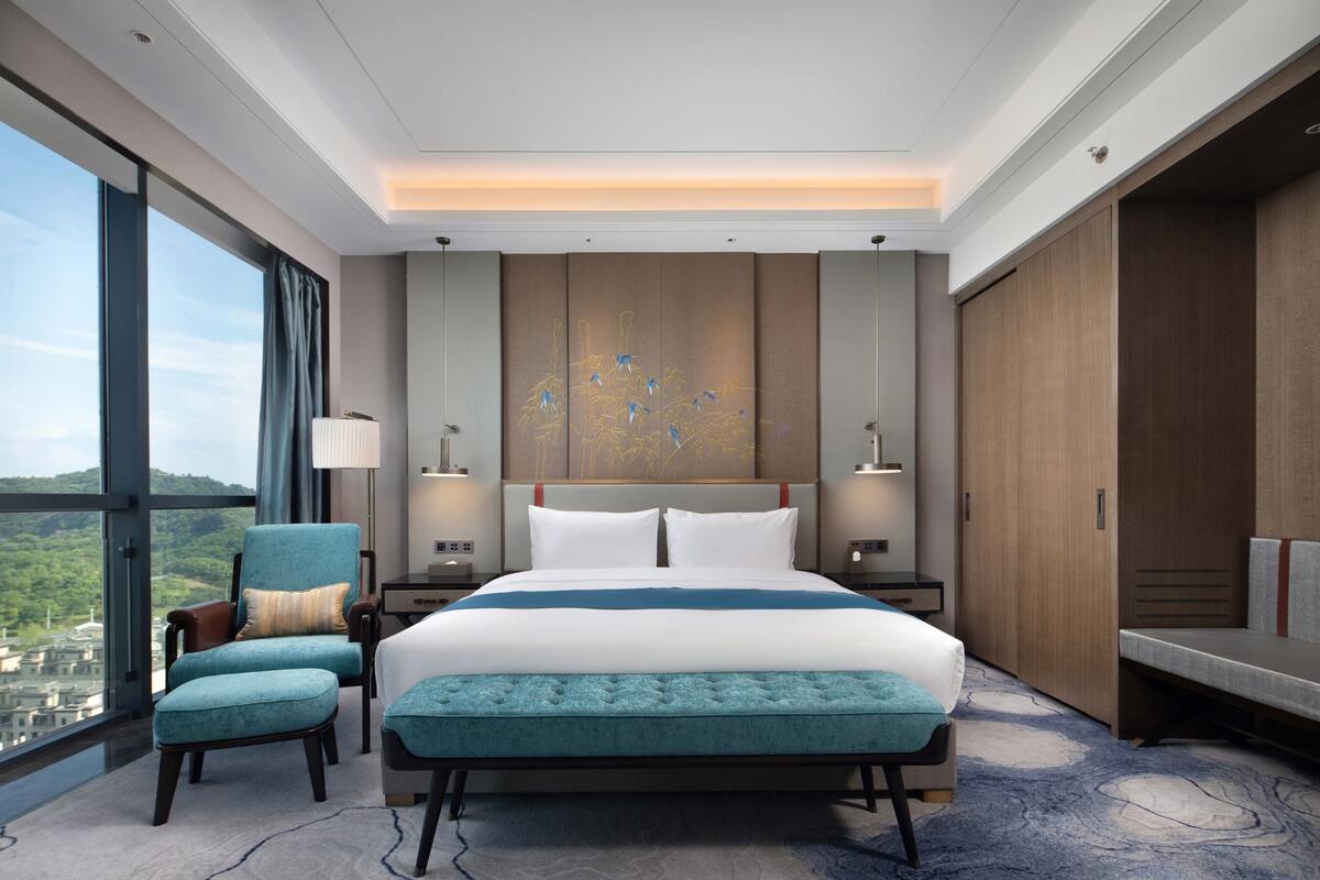 Una habitación de hotel moderna con una cama grande, una zona de estar con un banco, vistas a la ciudad y un armario.