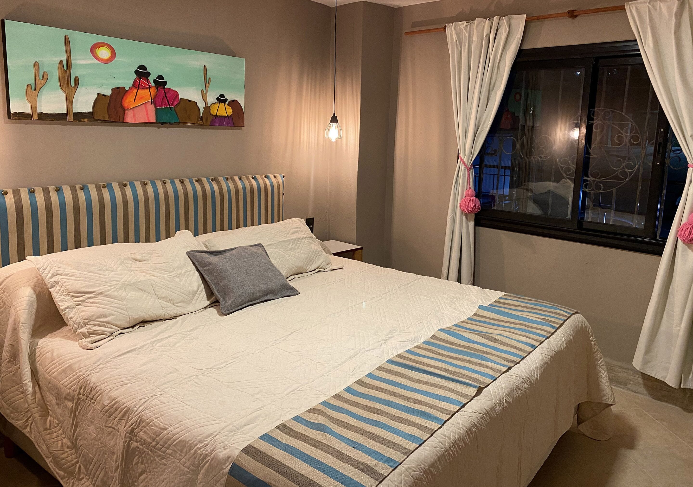 A hotel room with a bed, a painting on the wall, a window with curtains, and a radiator above the bed.