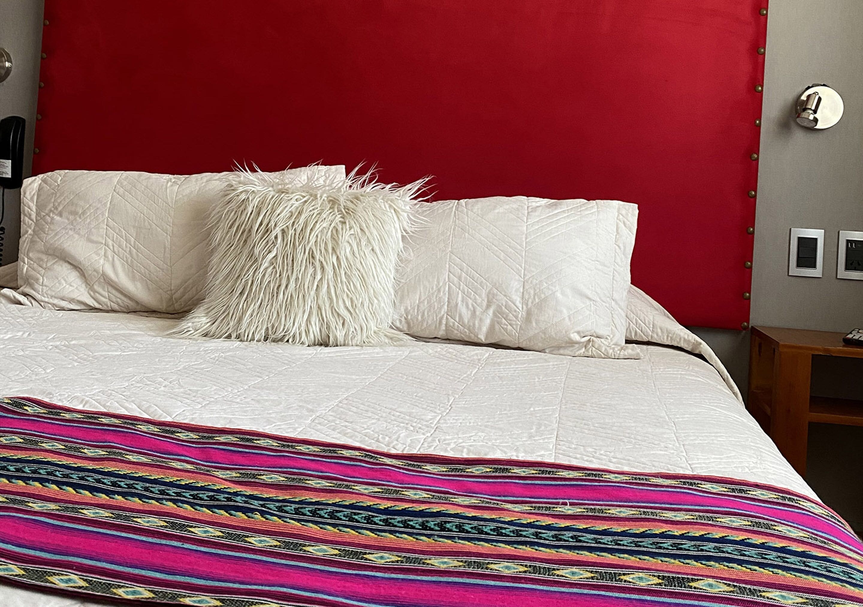 A bed with white bedding and a colorful blanket, a red headboard, and a small bedside table with a lamp.
