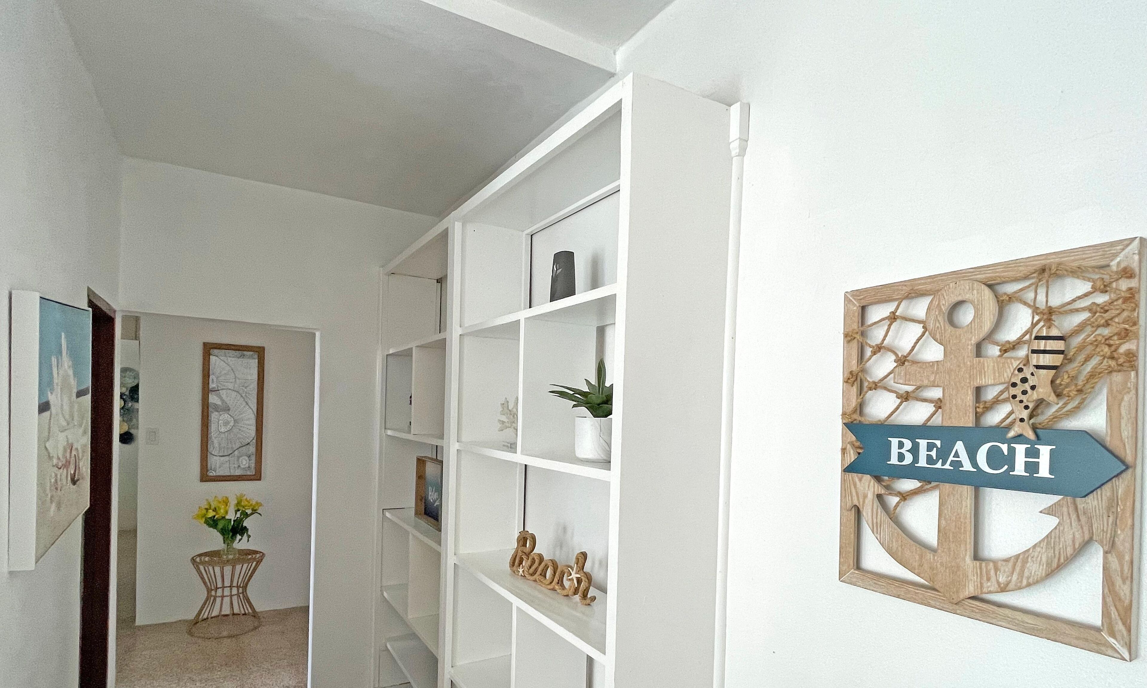 Un couloir avec des étagères blanches, une petite table sur laquelle est posé un vase de fleurs et une décoration murale avec le mot « PLAGE ».