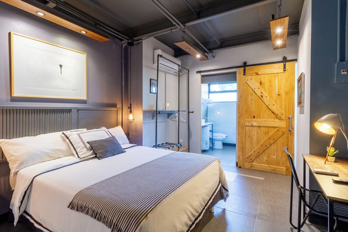A modern hotel room with a large bed, a wooden door, a desk with a lamp, and a framed picture on the wall.