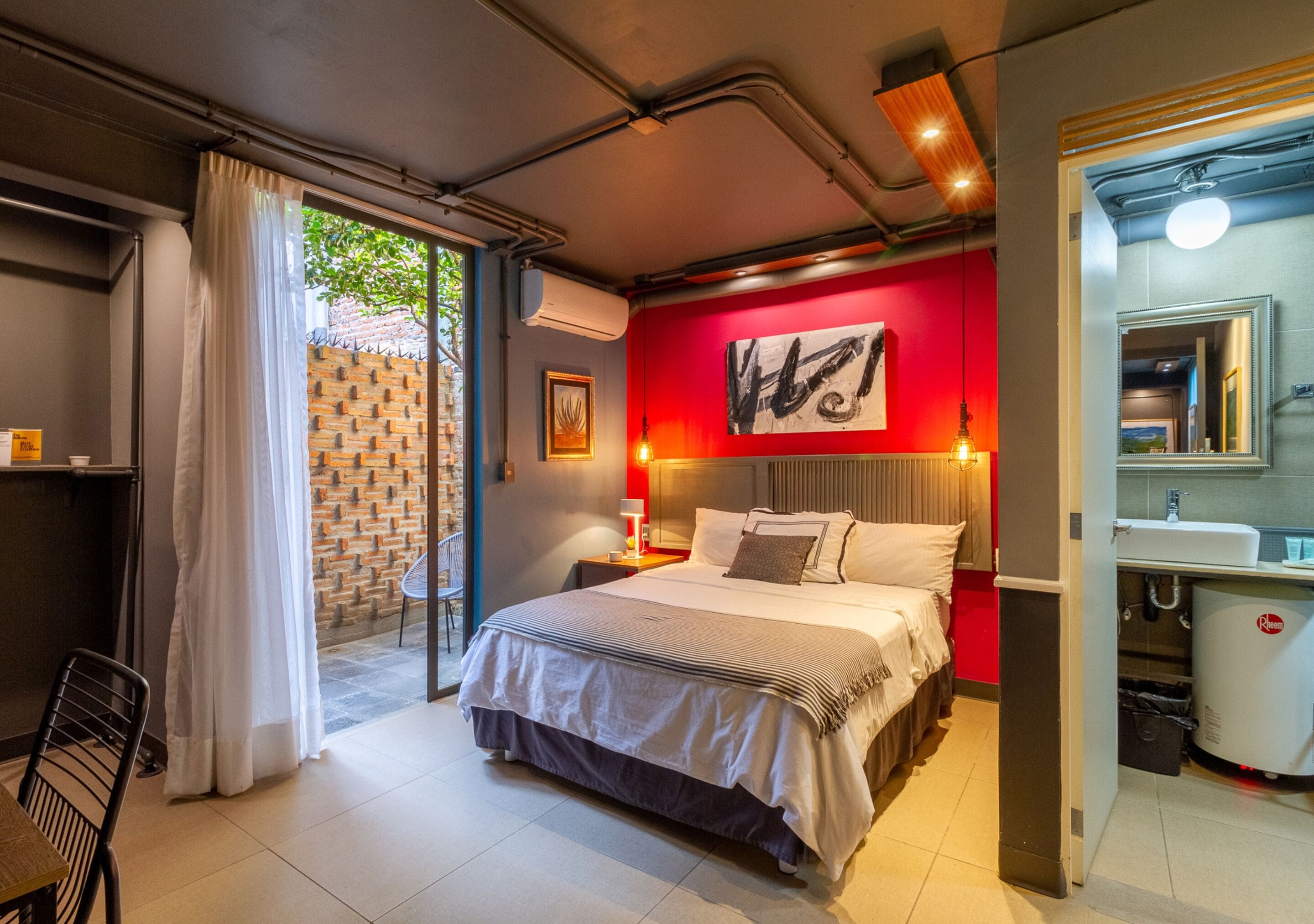Une chambre moderne avec un grand lit, un mur d’accent rouge, une salle de bain avec un lavabo et un miroir, et une vue sur l’extérieur grâce à une porte vitrée.