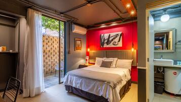 Une chambre moderne avec un grand lit, un mur d’accent rouge, une salle de bain avec un lavabo et un miroir, et une vue sur l’extérieur grâce à une porte vitrée.