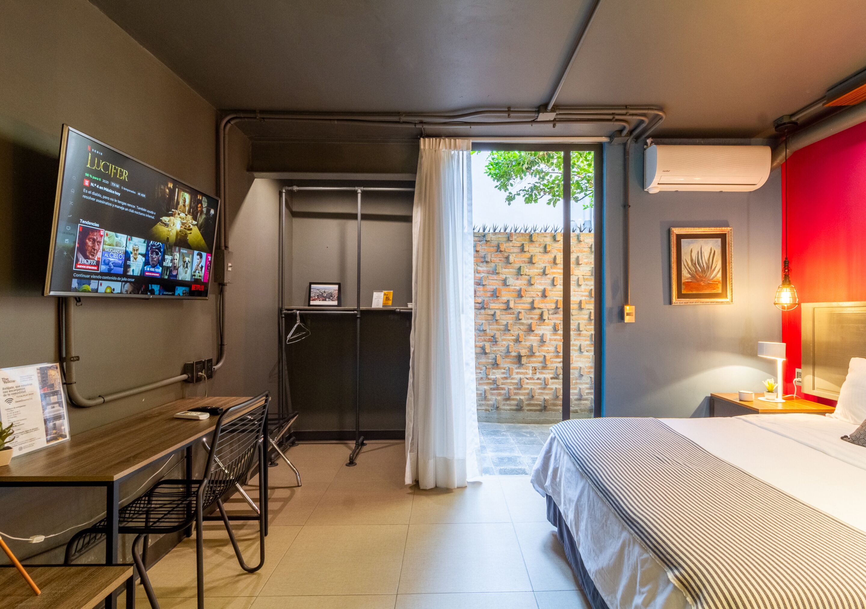 A hotel room with a bed, a desk with a chair, a TV mounted on the wall, and a window with curtains.