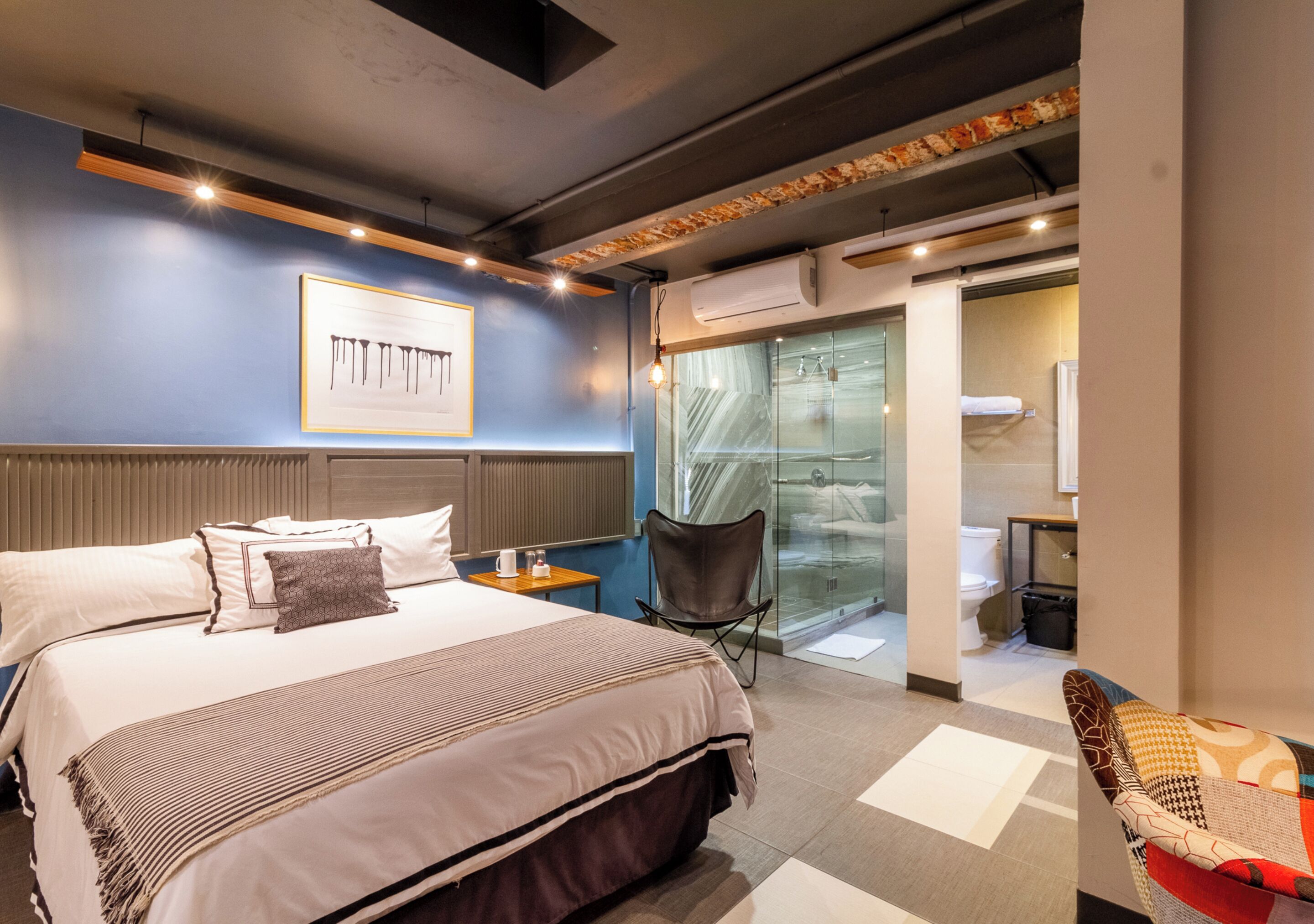 Une chambre d’hôtel moderne avec un grand lit, un coin salon comprenant un fauteuil et une petite table, ainsi qu’une porte vitrée donnant accès à la salle de bain.