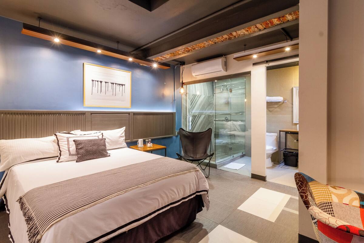 Une chambre d’hôtel moderne avec un grand lit, un coin salon comprenant un fauteuil et une petite table, ainsi qu’une porte vitrée donnant accès à la salle de bain.