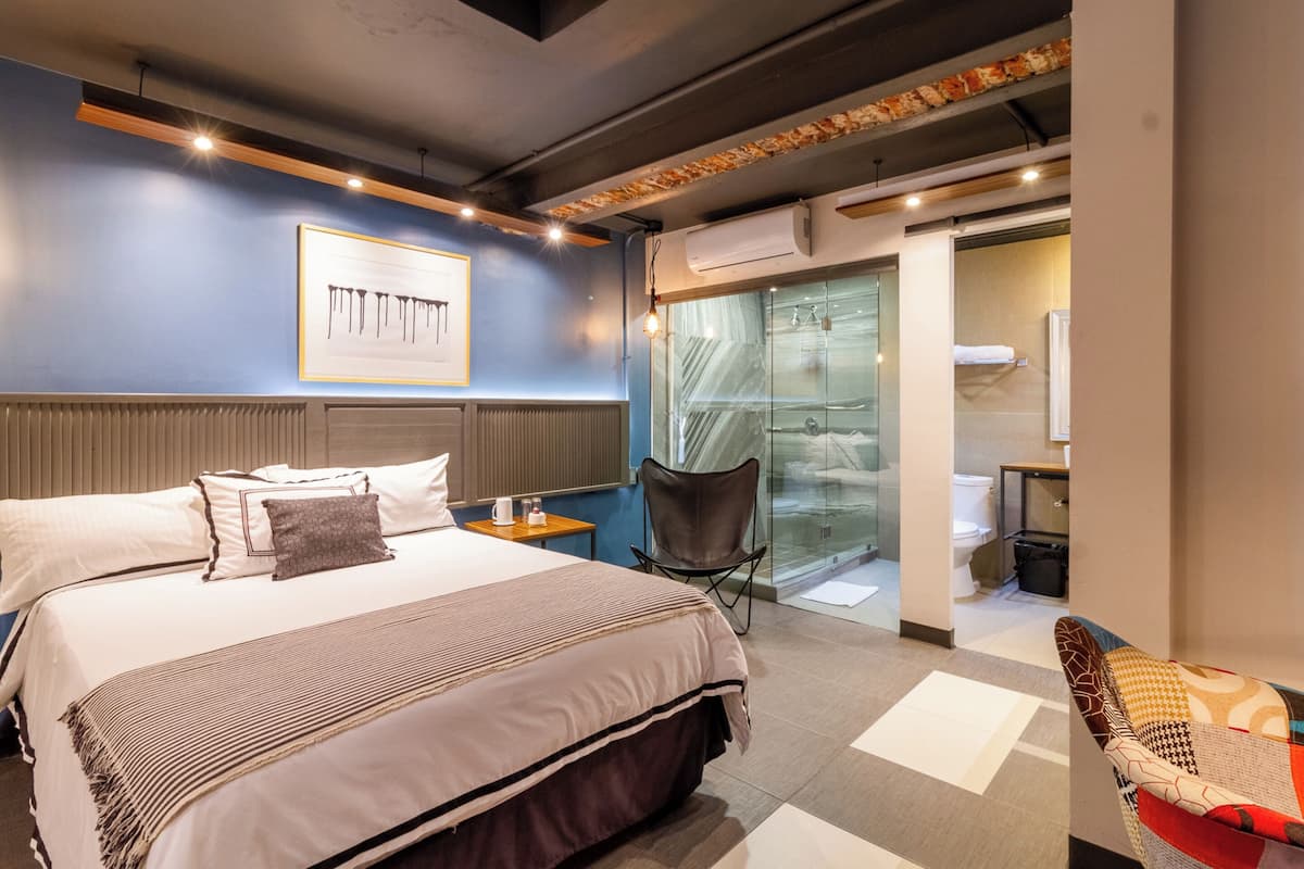 Una habitación de hotel moderna con una cama grande, una zona de estar con una silla, una mesita y una puerta de vidrio que da al baño.