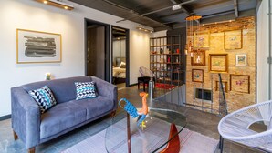 Een moderne woonkamer met een grijze bank, een glazen salontafel en een witte stoel. Er staat een boekenkast met ingelijste foto's en een kleine haanbeeldje.