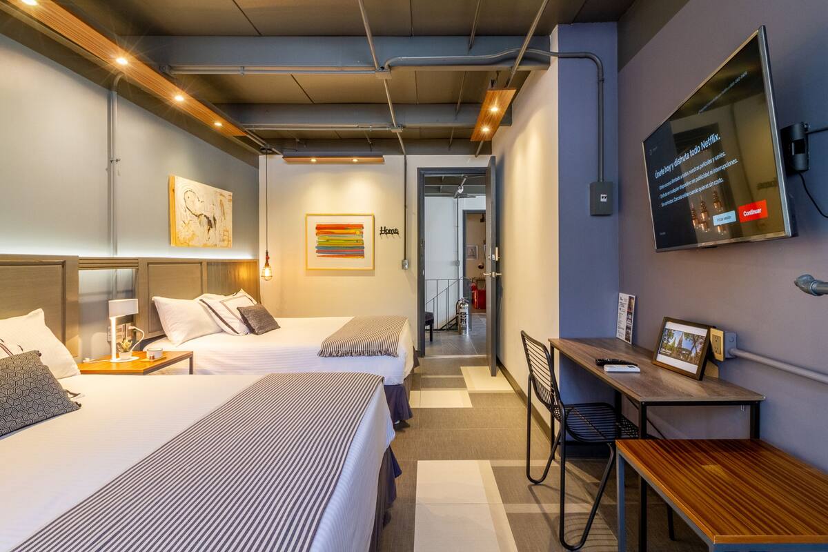 Une chambre d’hôtel moderne avec deux lits, un bureau, une chaise, une télévision et une salle de bain visible au fond.