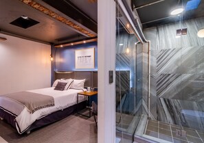 Una habitación de hotel moderna con cama, mesita de noche y zona de ducha con mampara de vidrio y baldosas de mármol.