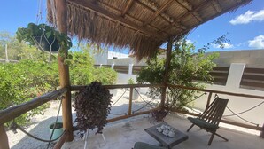 Un espacio de estar al aire libre cubierto con techo de paja, rodeado de follaje y vistas de edificios al fondo.
